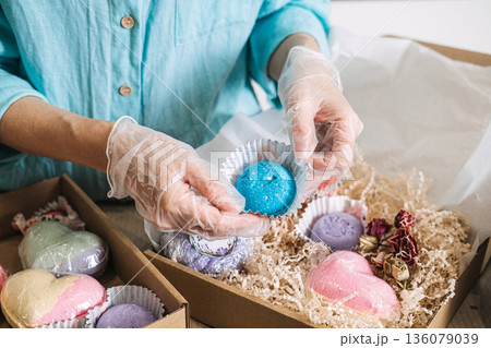 hands place blue bath bomb into cardboard gift box filled with shredded paper and various colorful cosmetic products. ceramide bath bomb, probiotic soak, lipid repair, skin longevity. 136079039