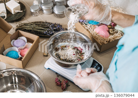Gloved hands pour dried flowers and seeds into metal bowl on digital scale surrounded by gift boxes. trash beauty, fruit waste, seed bombs, upcycled botanicals. Gloved hands pour dried flowers and seeds into metal bowl on digital scale surrounded by gift boxes. trash beauty, fruit waste, seed bombs, upcycled botanicals. 136079044
