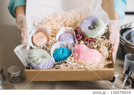 hands hold cardboard box filled with various colorful bath bombs and solid beauty products. solid shampoo bar, anhydrous conditioner, waterless skincare concentrate, plastic-free beauty bars. 136079059