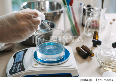 Gloved hand uses glass dropper to add liquid to beaker filled with clear fluid on digital scale. vegan PDRN, plant nucleotides, DNA repair, elasticity boost. 136079063