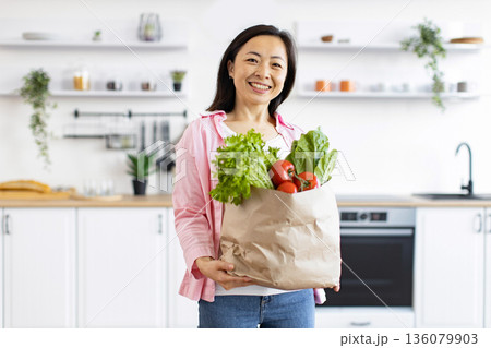 A smiling Asian woman holds a paper bag filled with fresh produce in her bright, modern kitchen A smiling Asian woman holds a paper bag filled with fresh produce in her bright, modern kitchen 136079903