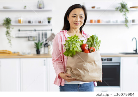 An Asian woman smiles while holding a bag of fresh produce in her bright, modern kitchen 136079904
