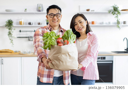 An Asian couple smiles while holding a bag of fresh produce in their bright, modern kitchen An Asian couple smiles while holding a bag of fresh produce in their bright, modern kitchen 136079905