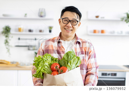 An Asian man smiles while holding a bag of fresh produce in his kitchen An Asian man smiles while holding a bag of fresh produce in his kitchen 136079932