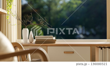 Books and dried branch in vase on wooden table with armchair aside wide window with nature scenery. Books and dried branch in vase on wooden table with armchair aside wide window with nature scenery. 136080321