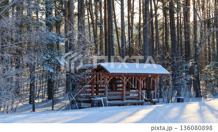 Wooden gazebo covered in snow within a serene winter landscape. Forest background with pine trees, snow covered ground, and long shadows Wooden gazebo covered in snow within a serene winter landscape. Forest background with pine trees, snow covered ground, and long shadows 136080898