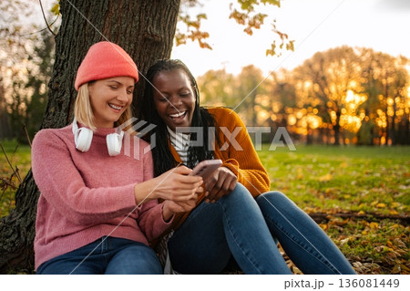 Diverse women friends laughing watching phone in autumn park Diverse women friends laughing watching phone in autumn park 136081449