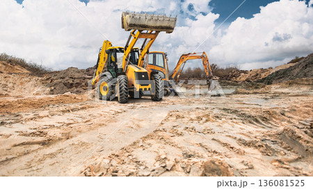 A loader and mini excavator are working in a construction quarry. Earthen embankments are visible, and the sky is partly cloudy. Work at the construction site continues 136081525