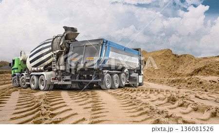 A large dump truck and concrete mixer truck at a construction site on a cloudy day. A large pile of sand is visible in the background A large dump truck and concrete mixer truck at a construction site on a cloudy day. A large pile of sand is visible in the background 136081526