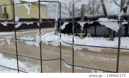 Horizontal wire fence blanketed in snow near yellow house and dark shed Horizontal wire fence blanketed in snow near yellow house and dark shed 136082177