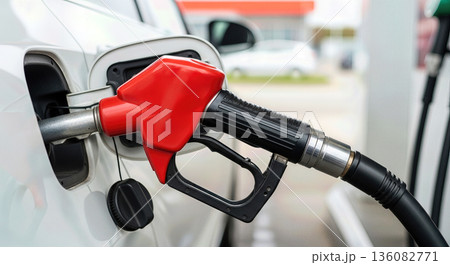 Close up of red fuel nozzle inserted into white car tank during refueling process at petrol station for transportation energy concept Close up of red fuel nozzle inserted into white car tank during refueling process at petrol station for transportation energy concept 136082771