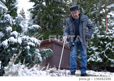 Elderly man walking with cane on paved yard after snowfall wearing winter jacket and hat outdoors calm winter environment. 136082886