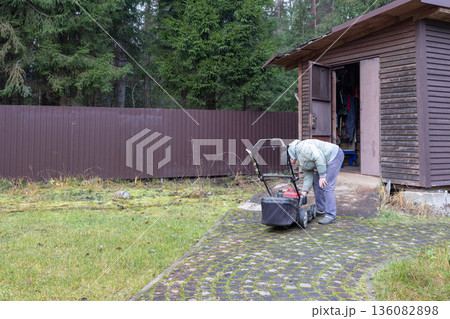 Man inspects red lawnmower near wooden house wearing puffy jacket beanie crouched in outdoor setting. 136082898
