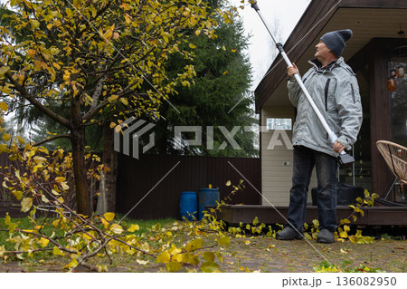 Male homeowner performing tree pruning with power tool, fall season, routine landscape maintenance, practical suburban lifestyle activity. Male homeowner performing tree pruning with power tool, fall season, routine landscape maintenance, practical suburban lifestyle activity. 136082950
