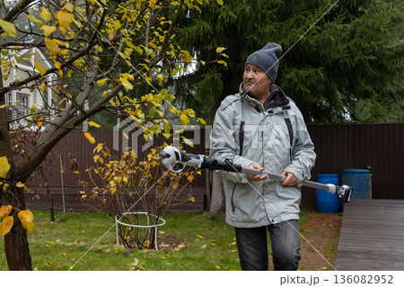 Adult man trimming branches using electric pole pruner, autumn foliage, seasonal garden care, outdoor work in private residential yard. Adult man trimming branches using electric pole pruner, autumn foliage, seasonal garden care, outdoor work in private residential yard. 136082952