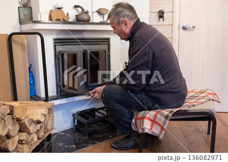Elderly man lights fireplace into indoor fireplace preparing home heating during cold season domestic routine. Elderly man lights fireplace into indoor fireplace preparing home heating during cold season domestic routine. 136082971