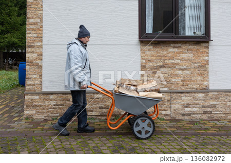 Adult man pushing wheelbarrow loaded with firewood along stone path in yard wearing warm jacket and hat. 136082972