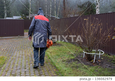 Worker removing fallen leaves with electric blower, damp weather, everyday maintenance task, suburban environment, realistic lifestyle scene. Worker removing fallen leaves with electric blower, damp weather, everyday maintenance task, suburban environment, realistic lifestyle scene. 136082979