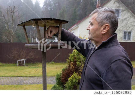 Man refilling bird feeder with seeds in autumn garden among trees during cold season. 136082989