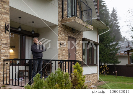 Mature man standing on balcony holding cigarette calm thoughtful mood, casual clothing in residential exterior. Everyday lifestyle moment. Mature man standing on balcony holding cigarette calm thoughtful mood, casual clothing in residential exterior. Everyday lifestyle moment. 136083015