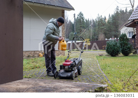 Man refuels red lawnmower using yellow fuel container. Concept yard work in residential area. Man refuels red lawnmower using yellow fuel container. Concept yard work in residential area. 136083031