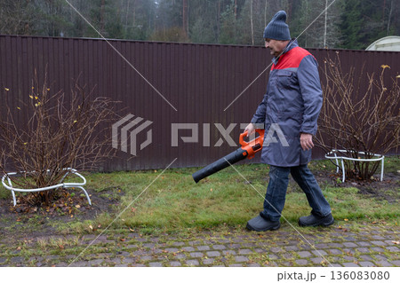 Worker removing fallen leaves with electric blower, damp weather, everyday maintenance task, suburban environment, realistic lifestyle scene.  136083080