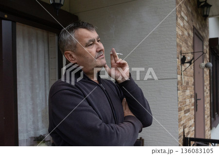 Mature man standing on balcony holding cigarette calm thoughtful mood, casual clothing in residential exterior. Everyday lifestyle moment. 136083105