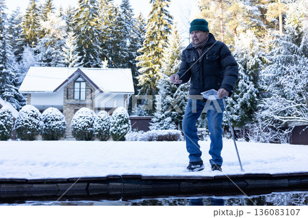 Male person near water pond throwing food to fish on snow covered landscape. Peaceful lifestyle moment on natural light scene. Male person near water pond throwing food to fish on snow covered landscape. Peaceful lifestyle moment on natural light scene. 136083107