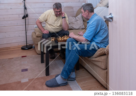 Elderly friends concentrating on chess match in living room in casual clothing. Thoughtful mood and indoor recreation scene. Elderly friends concentrating on chess match in living room in casual clothing. Thoughtful mood and indoor recreation scene. 136083118