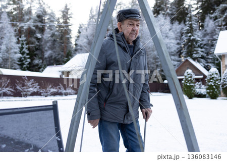 Elderly man walking with cane on paved yard after snowfall wearing winter jacket and hat outdoors calm winter environment. Elderly man walking with cane on paved yard after snowfall wearing winter jacket and hat outdoors calm winter environment. 136083146