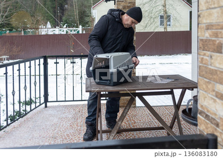 Adult man in warm winter clothing adjusting plastic pet carrier on outdoor terrace with snow around. 136083180