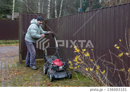 Man mows lawn with red lawnmower in front of house, greenery around, overcast day. Garden maintenance task in overcast day 136083181