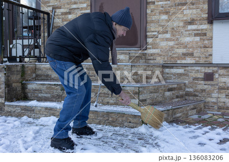 Male person removing snow using broom near house entrance. Snowy yard environment, routine domestic activity in daylight scene. 136083206