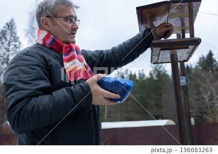 Man refilling bird feeder with seeds in snowy garden wearing jacket and scarf among trees during cold season. Man refilling bird feeder with seeds in snowy garden wearing jacket and scarf among trees during cold season. 136083263