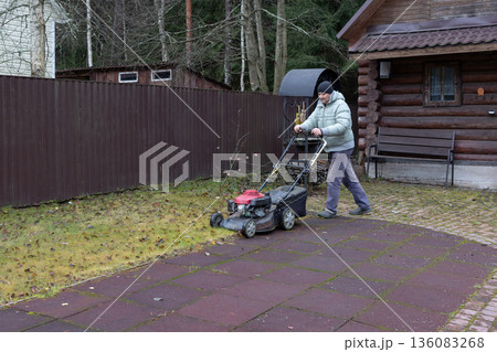 Man uses lawnmower to trim grass in front of residential home and lush greenery. Calm outdoor setting. Man uses lawnmower to trim grass in front of residential home and lush greenery. Calm outdoor setting. 136083268