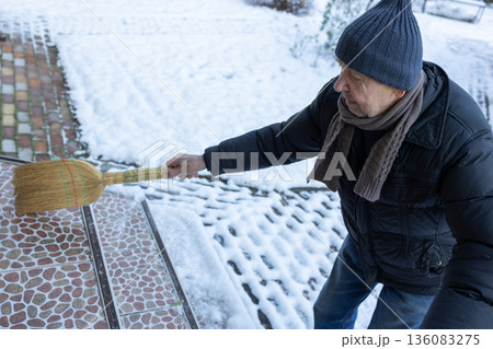 Male person removing snow using broom near house entrance. Snowy yard environment, routine domestic activity in daylight scene. 136083275