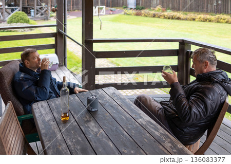 Two adult men with whiskey glasses at wooden table on outdoor patio with brick wall wearing jackets in casual setting. Two adult men with whiskey glasses at wooden table on outdoor patio with brick wall wearing jackets in casual setting. 136083377
