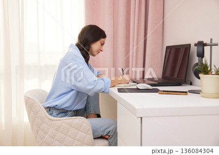 Woman writing in a notebook while working or studying with a laptop at a desk Woman writing in a notebook while working or studying with a laptop at a desk 136084304