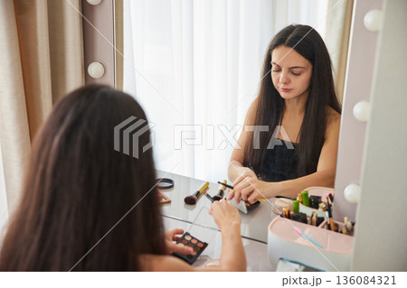 Woman holding cosmetic tube and brush, preparing makeup routine in front of mirror 136084321