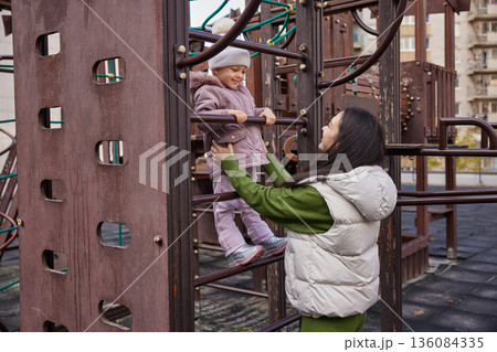 Mother assisting child on monkey bars at a playground, sharing a happy moment Mother assisting child on monkey bars at a playground, sharing a happy moment 136084335