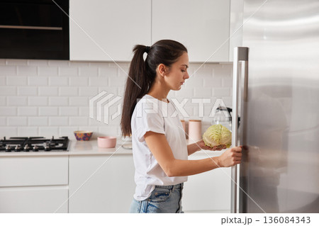 Young woman putting healthy organic vegetable into modern kitchen fridge for storage Young woman putting healthy organic vegetable into modern kitchen fridge for storage 136084343