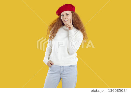 Beautiful young happy woman with long wavy hair in red beret and white sweater touches her chin, smiles and looks at camera. 136085084