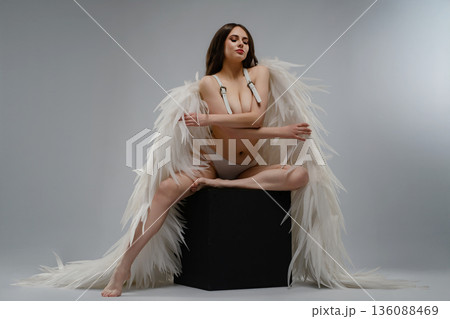 Ethereal woman in white angel wings posing on a black cube against a grey background 136088469