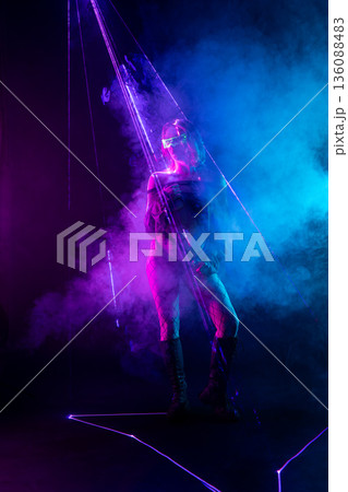 Cyberpunk woman in futuristic glasses stands amidst neon purple laser beams and blue smoke. Cyberpunk woman in futuristic glasses stands amidst neon purple laser beams and blue smoke. 136088483