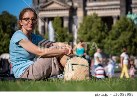 Berlin, germany, august 11, 2023. Woman sitting on the green grass, relaxing and looking at the camera, with the berlin cathedral in the blurry background 136088989