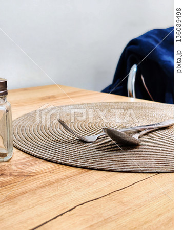 A round placemat is placed on a wooden table. A spoon and a fork are resting on the placemat next to a small glass jar. A chair is in the background 136089498