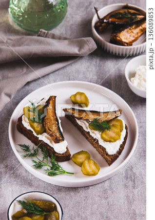 Rye bread toasts with cream cheese, sprats and pickles on a plate on the table vertical view Rye bread toasts with cream cheese, sprats and pickles on a plate on the table vertical view 136089853