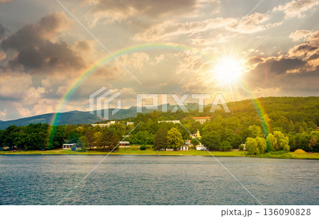 scenic landscape with shore of a lake in summer at sunset. beautiful scenery of body of water in evening light. distant forested mountain. countryside place for recreation or camping under rainbow 136090828