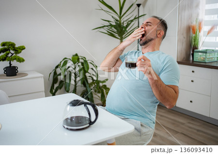 Sleepy mid adult man yawning holding coffee at home Sleepy mid adult man yawning holding coffee at home 136093410