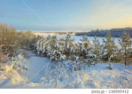 A snow-covered landscape with trees and a frozen river A snow-covered landscape with trees and a frozen river 136095412